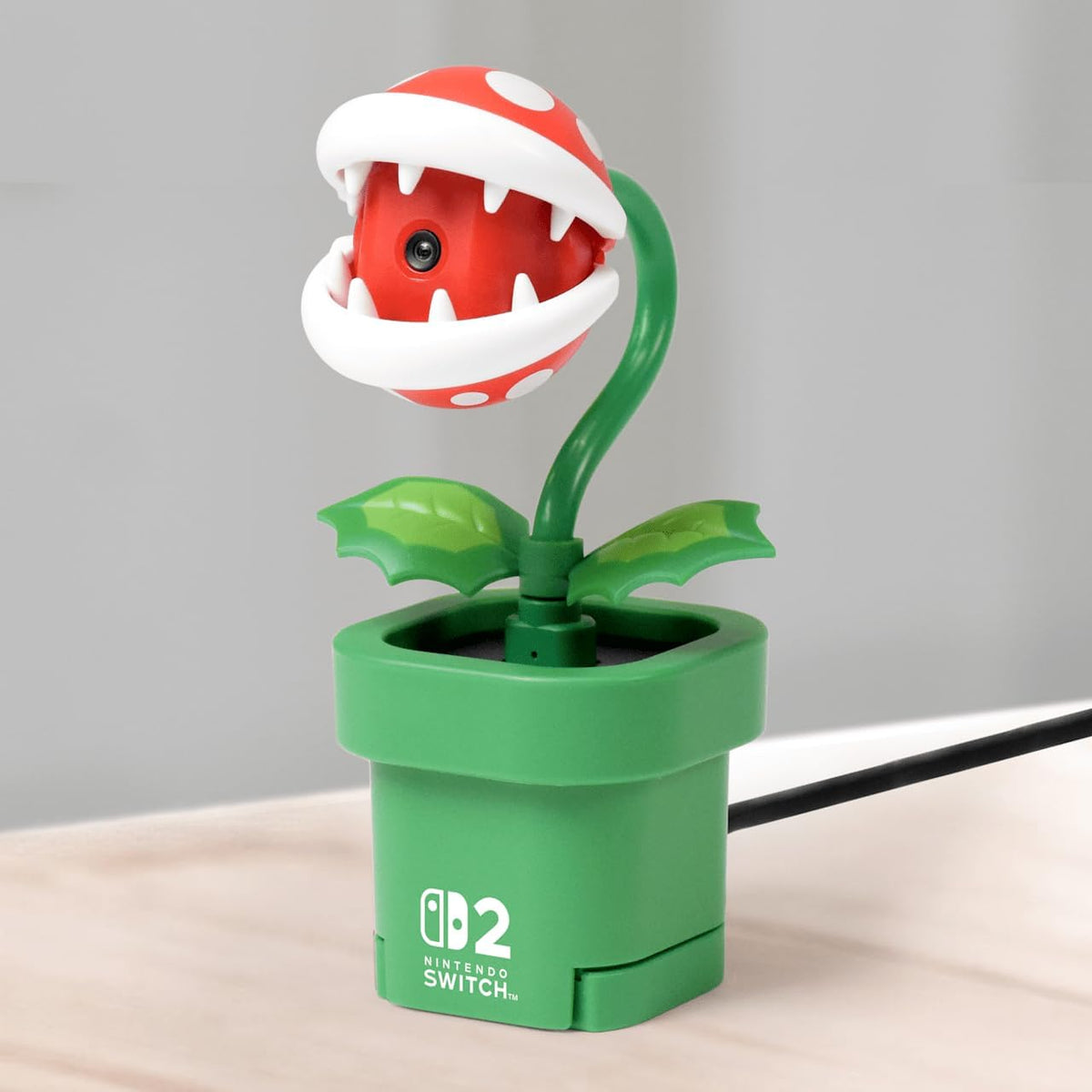 HORI Piranha Plant Camera for Nintendo Switch 2 displayed on its green Warp Pipe stand, showing the red and white plant design and connected USB-C cable.