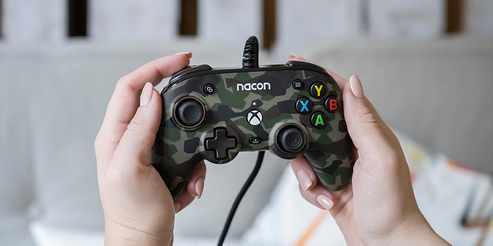 Nacon Pro Compact Forest Camo wired Xbox controller held in both hands, demonstrating its compact ergonomic shape, analogue stick positioning and gaming button layout.