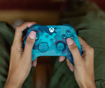 Microsoft Xbox Mineral Camo Wireless Controller held in both hands, showing its colourful blue camouflage design, comfortable ergonomic grips and gaming button layout.